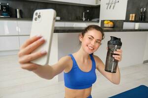 retrato de deportista toma selfie con agua botella en vivo habitación, sostiene teléfono inteligente y poses para foto mientras haciendo aptitud rutina de ejercicio