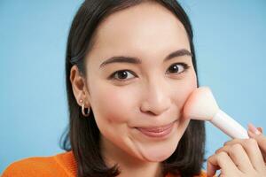 Makeup and beauty. Cute asian woman using cosmetic brush, puts on blush or highlighter, stands over blue background photo