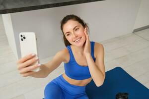 Image of beautiful, atheltic girl sits on yoga mat at home with smartphone, takes selfie and workout, does fitness exercises photo