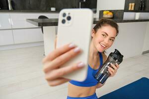 Portrait of sportswoman takes selfie with water bottle in living room, holds smartphone and poses for photo while doing fitness workout