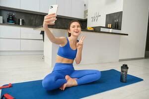 Active young woman, vlogger does sports, records her workout training from home on smartphone camera, posing for selfie inside her house, sits on rubber yoga mat in blue leggings and sportsbra photo