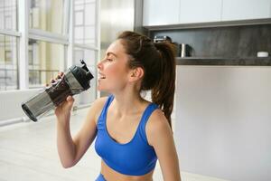 Portrait of sportswoman drinking water during workout session in living room, does her fitness training exercises at home photo