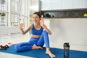 Image of beautiful, atheltic girl sits on yoga mat at home with smartphone, takes selfie and workout, does fitness exercises photo
