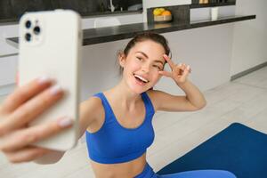 cerca arriba retrato de hermosa morena deportista, tomando selfie en teléfono inteligente, posando en aptitud estera con móvil teléfono, registros su aptitud ejercicios, rutina de ejercicio formación foto