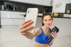 retrato de joven aptitud chica, toma selfie en teléfono inteligente con agua botella, bebidas agua durante ejercicio, formación a hogar, registros Deportes vlog foto