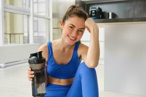 Smiling fitness girl drinks water in the middle of training session, smiling and looking happy, workout from home in blue sportsbra photo