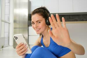 Happy fitness instructor at home, does workout with headphones and smartphone, listens to music and doing training exercises photo