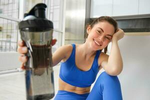 Young woman working out from home, drinking water during her sport training session in living room, sitting on yoga mat photo