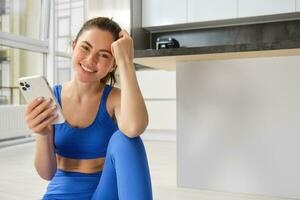 Young woman with smartphone does workout at home, using mobile phone app for sports training indoors, wears blue sportsbra and leggings photo