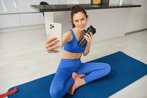 Portrait of woman doing sports, takes selfie on smartphone, fitness instructor records her exercises, stays hydrated, drinks water during training session photo