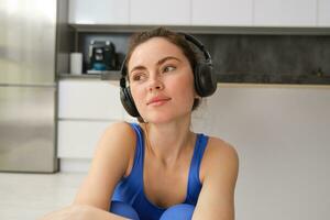 Beautiful brunette girl, doing fitness workout, sitting in living room in wireless headphones, listening to music and doing sports photo