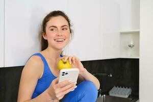 Portrait of stylish, young fitness woman, eating an apple and using mobile phone, holding smartphone, wearing sportswear photo