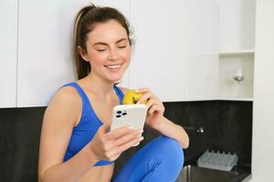 sano estilo de vida y tecnología. joven hermosa chica, aptitud instructor, mirando a teléfono inteligente, leyendo en móvil teléfono y comiendo un manzana, sonriente felizmente foto