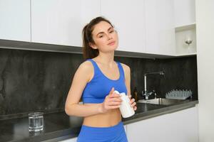 Image of young brunette woman with slim, fit body, holding dietary supplements, vitamin B, fish oil, standing in kitchen photo