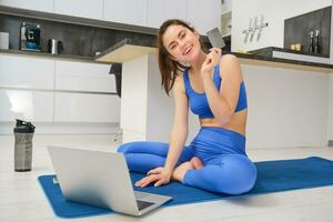 Image of smiling young woman, holding credit card, sitting on fitness mat with laptop, buying sport app subscription, workout video tutorials, doing exercises at home photo