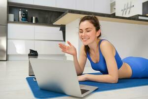 Image of young fitness girl talking online on laptop, video chats, laying on floor rubber mat, doing exercises, discuss workout training, yoga from home photo