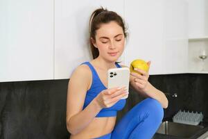 Portrait of fitness woman, sitting in kitchen with smartphone, scrolling social media and eating an apple, having a healthy snack between workout, wearing sports activewear photo