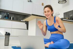 Portrait of fit and healthy, female fitness instructor sitting on floor at home, talking to laptop, teaching online class yoga or workout training photo