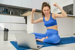 Image of excited young woman showing muscles, flexing biceps and looking at laptop screen, workout from home, fitness instructor teaching remotely, giving online yoga lesson photo