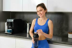 Portrait of fit and healthy smiling woman, wearing fitness sportswear, standing in kitchen with water bottle and looking happy photo