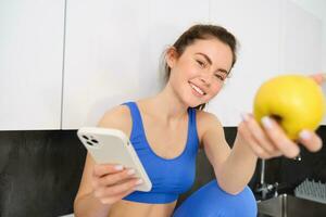 Portrait of beautiful, smiling young fitness woman, offering you an apple, eating healthy snack, holding a fruit, sitting in kitchen photo