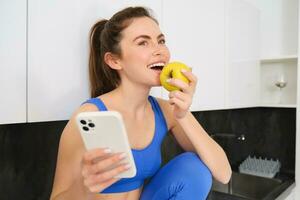 retrato de elegante, joven aptitud mujer, comiendo un manzana y utilizando móvil teléfono, participación teléfono inteligente, vistiendo ropa de deporte foto