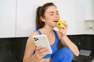 Image of stylish sportswoman, young fitness instructor sitting in kitchen and eating an apple, holding smartphone, using social media app on mobile phone photo