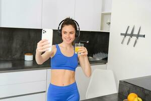 Portrait of young lifestyle, fitness blogger, woman with orange juice, takes selfie on smartphone camera, posing in activewear and wireless headphones photo