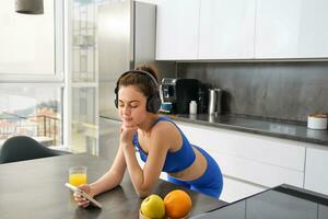 Portrait of young modern woman, beautiful girl in fitness clothes, standing in kitchen, watching yoga video class on smartphone, listening in wireless headphones, drinking juice photo