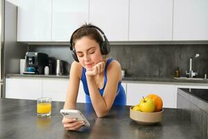 imagen de feliz, elegante joven Deportes mujer, en pie en cocina y Bebiendo naranja jugo, escuchando música en auriculares, utilizando teléfono inteligente aplicación foto