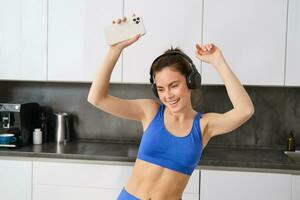 retrato de despreocupado deportista, bailando en cocina con teléfono inteligente, escuchando música en auriculares, sensación energizado y emocionado después rutina de ejercicio foto