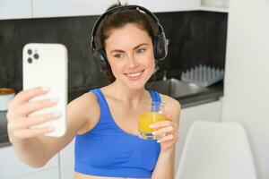 cerca arriba retrato de sonriente morena mujer, aptitud niña toma selfie para su social medios de comunicación, usa inalámbrico auriculares, sostiene vaso de naranja jugo, soportes en cocina en ropa de deporte foto