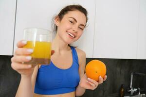 Portrait of smiling fitness woman, offering orange juice, holding fruit and a glass in hands, posing in kitchen in activewear photo