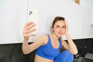 retrato de hermosa deportista, vistiendo sujetador deportivo y polainas, sentado en cocina y tomando autofoto, posando para foto en teléfono inteligente aplicación