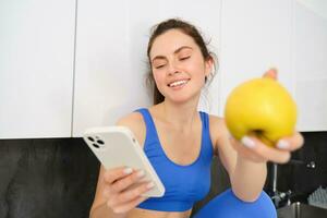 retrato de sonriente, contento aptitud chica, sentado con un manzana, riendo terminado algo en teléfono inteligente, hojeada social medios de comunicación en móvil teléfono aplicación foto