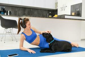 Portrait of happy young woman, tried to workout at home, laying on yoga mat in activewear, playing with her dog, listening music in wireless headphones photo