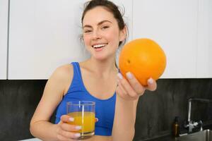 Close up portrait of sportswoman, fitness girl holding glass of fresh juice and an orange in hands, smiling at camera, standing in kitchen photo