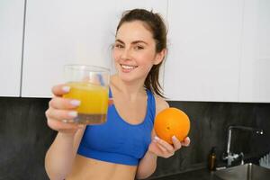 retrato de sonriente, hermosa joven mujer, ofrecimiento vaso de Fresco jugo, participación un naranja y mirando feliz, aptitud instructor dando usted un bebida foto