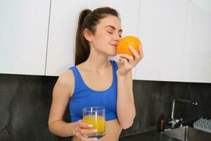 Portrait of fitness instructor, smiling woman, enjoys smell of fresh fruit, drinking orange juice and looking happy, standing in kitchen in activewear photo