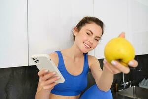 retrato de hermoso, sonriente joven aptitud mujer, ofrecimiento usted un manzana, comiendo sano bocadillo, participación un fruta, sentado en cocina foto