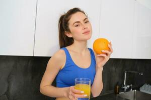 Indoor shot of young brunette woman in sportswear, drinking orange juice, holding fruit in hand, posing in kitchen photo