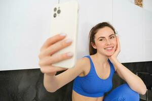 Portrait of fitness girl posing for photo, taking selfie on smartphone app, sitting in kitchen, wearing activewear photo