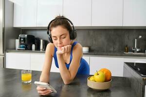 Portrait of young fitness woman with headphones, drinking orange juice in kitchen and using smartphone, listening music, getting ready for workout gym photo