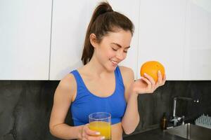 bienestar y deporte. joven sonriente nutricionista, aptitud niña participación naranja y Fresco jugo, Bebiendo eso desde vaso y mirando feliz, posando en sujetador deportivo y polainas, en pie en cocina foto