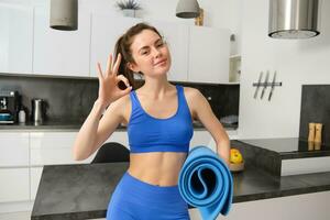 Smiling brunette woman, fitness instructor approves yoga app, mindfulness application, workout at home, standing in living room, showing ok gesture and holding rubber mat photo