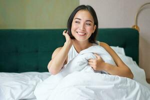 Close up of happy beautiful asian woman, waking up in bed and enjoys morning, looking outside window with sleepy smile on her face, hugging duvet photo