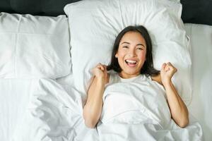 Top view of enthusiastic asian girl cheering, lying in her bed and shouting with joy and excitement, triumphing and smiling, enjoy staying in her bedroom photo