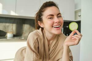 Food and lifestyle concept. Beautiful woman cooking in the kitchen, holding raw zucchini and smiling, preparing healthy vegetarian meal, making salad, looking happy photo