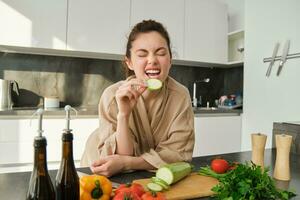 Portrait of happy girl eating vegetables while making meal, cooking healthy vegetarian food in kitchen, holding zucchini, posing in bathrobe photo