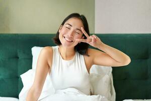 Positive asian woman lying in bed, showing peace sign, enjoys happy morning, waking up upbeat, staying in her bedroom photo